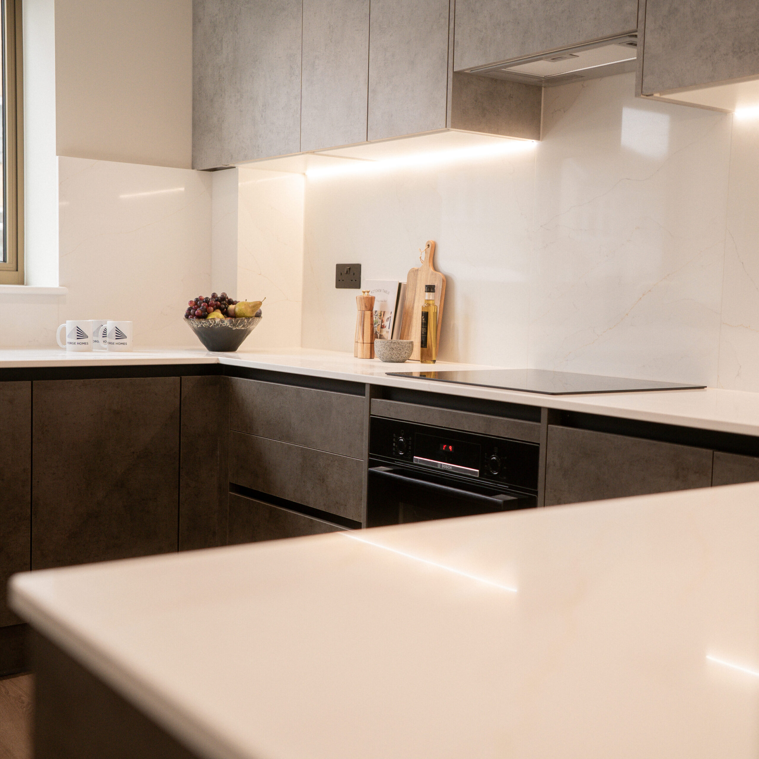 Light slate grey top cabinets with white marble splashback and countertops. Led lights underneath the grey top cabinets. Decorative items on top of worktop, with mugs, fruit and countertop, oils, and other cooking utensils