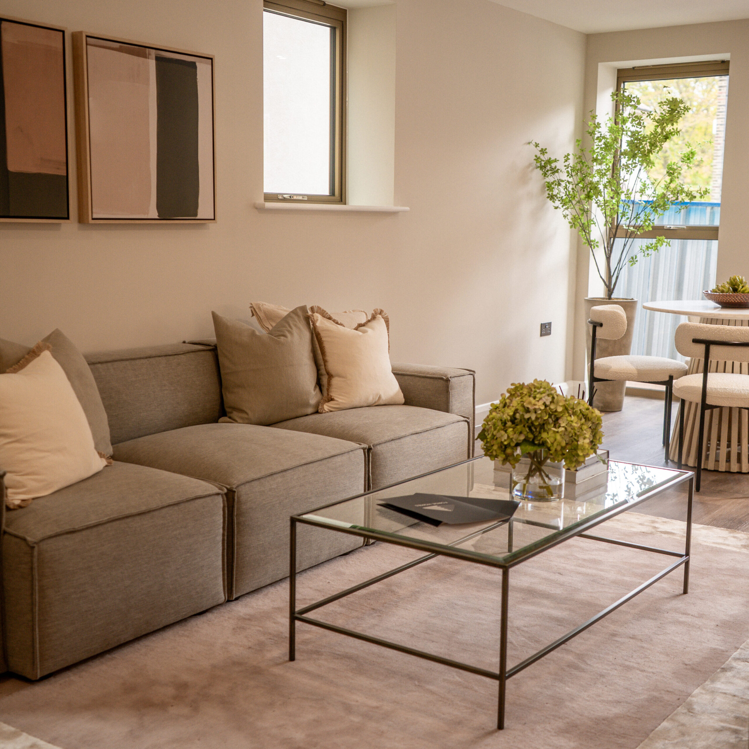 Living room, with gold exterior windows and grey sofas. Glass coffee table with more decorative pieces on top. Large purple tone rug underneath the sofa and table. Decorative artificial tree in the corner with dining table