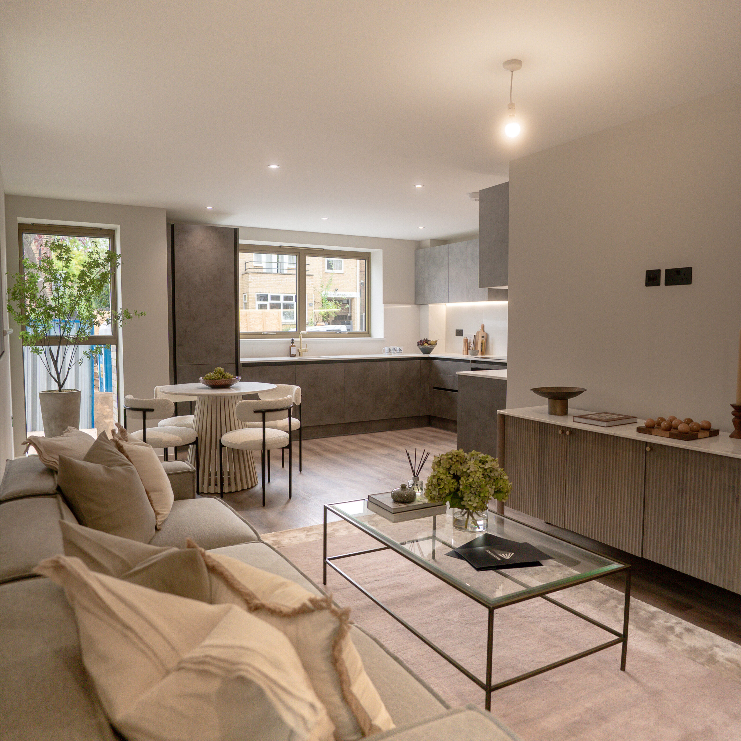 Living room, with gold exterior windows and grey sofas. Glass coffee table with more decorative pieces on top. Large purple tone rug underneath the sofa and table. Decorative artificial tree in the corner with dining table. Kitchen in the background with slate grey cabinets and marble countertops